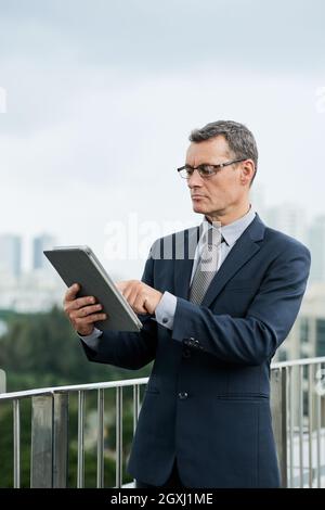Concentrato uomo d'affari caucasico maturo in abito scuro in piedi sul balcone e tablet digitale usin Foto Stock