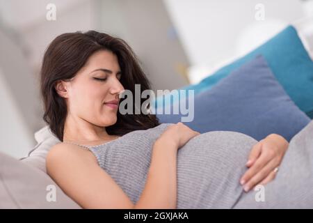 Felice giovane donna incinta mantenendo la mano sulla pancia mentre è seduto sul divano di casa Foto Stock