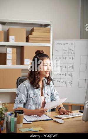 Positivo giovane amministratore di ufficio asiatico con capelli ricci seduti alla scrivania di legno e l'inserimento di dati nel computer Foto Stock
