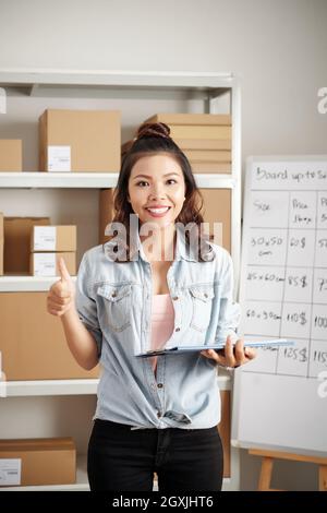 Ritratto di giovane operaio postale asiatico felice con capelli ricci che tengono cartella con documenti e mostrando pollice-su Foto Stock