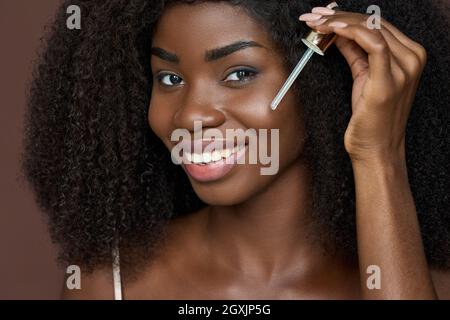 Sorridente giovane donna nera africana felice applicare olio di siero sulla pelle del viso. Foto Stock