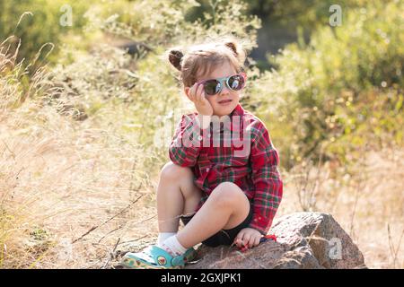 Piccola ragazza rossa sorridente in occhiali da sole e ponytail su uno sfondo rustico sfocato. Il bambino bello carino in una camicia marrone rossa si siede su una pietra grande Foto Stock