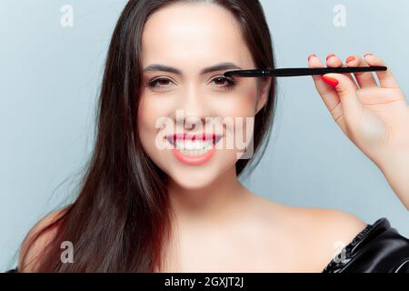 un giovane modello di brunetta tiene una spazzola per il trucco vicino al suo occhio e sorride ampiamente mostrando i suoi denti Foto Stock