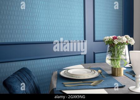 Tavolo per cena con piatti bianchi e matematica da tavolo blu, argenteria placcata oro e fiori in vaso contro parete blu Foto Stock