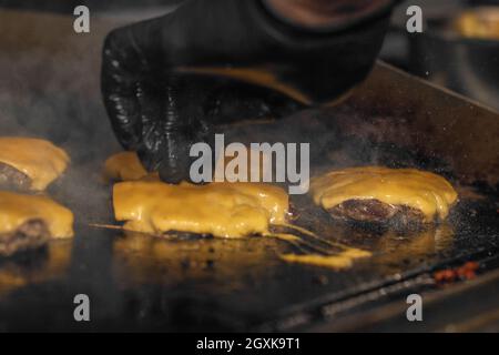 Processo di cottura degli cheeseburgers sul grill. Chef in guanti neri che cucinano un hamburger Foto Stock