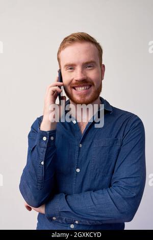 Ritratto di allegro giovane rosso-bearded manager in camicia blu scuro comunicando dal telefono cellulare Foto Stock