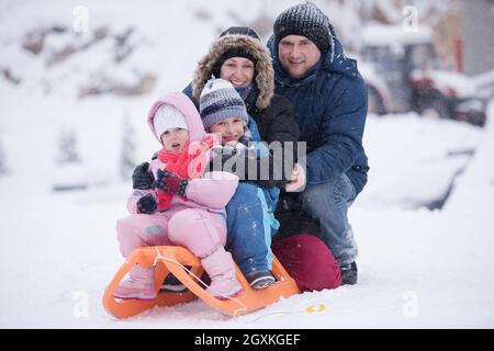 Felice giovani ritratto di famiglia per le vacanze invernali seduti slitta al paesaggio con un fresco e caduta di neve Foto Stock