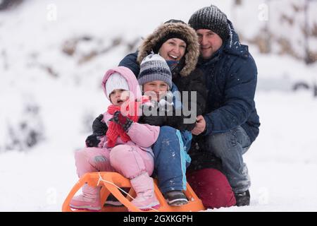 Felice giovani ritratto di famiglia per le vacanze invernali seduti slitta al paesaggio con un fresco e caduta di neve Foto Stock