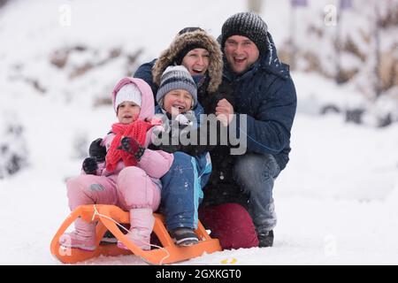 Felice giovani ritratto di famiglia per le vacanze invernali seduti slitta al paesaggio con un fresco e caduta di neve Foto Stock