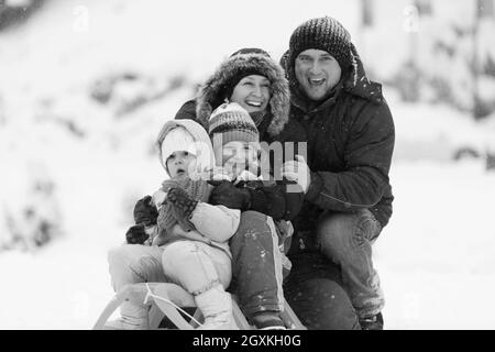 Felice giovani ritratto di famiglia per le vacanze invernali seduti slitta al paesaggio con un fresco e caduta di neve Foto Stock