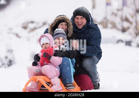 Felice giovani ritratto di famiglia per le vacanze invernali seduti slitta al paesaggio con un fresco e caduta di neve Foto Stock