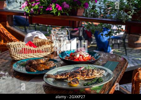 tavolo del server con un pranzo di tre portate preparato al momento sulla terrazza estiva della caffetteria. Trota alla griglia, patate alla griglia, pomodoro fresco, pepe e o Foto Stock