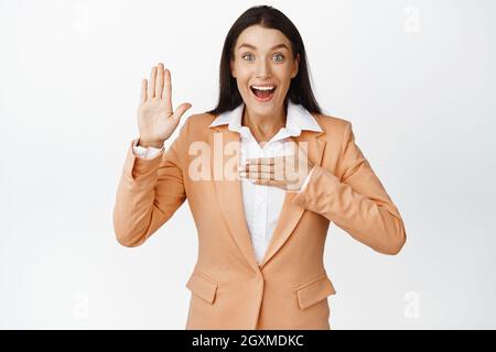 Donna corporativa eccitata che alza una mano e che osserva felice, facendo la promessa, levandosi in piedi nel vestito contro sfondo bianco Foto Stock