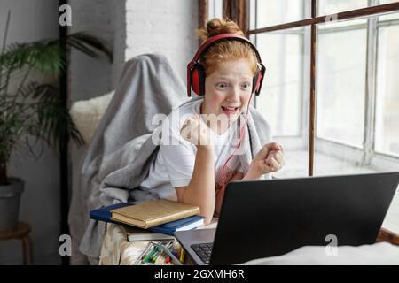 Adolescente ragazza impara da remoto. Il bambino ascolta una lezione online su un notebook. Grande finestra. Cuffie rosse. Foto Stock