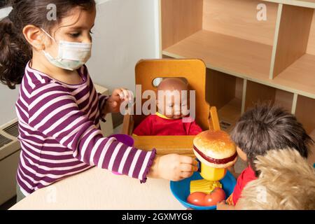 Istruzione Preschool 3-4 anni fingono di giocare in famiglia, bambola che allattano con cibo da gioco, indossare maschera per il viso per proteggere contro Covid-19i Foto Stock