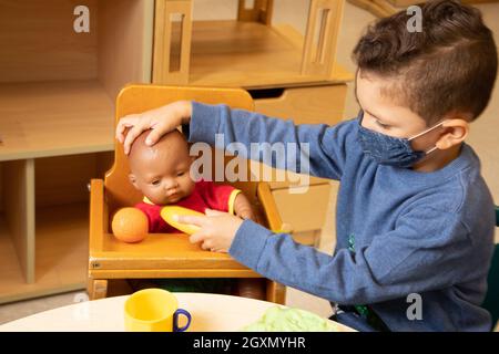 Istruzione Preschool 3-4 anni fingono di giocare in famiglia, ragazzo che allattano bambola con cibo da gioco, indossare maschera per il viso per proteggere contro Covid-19 Foto Stock