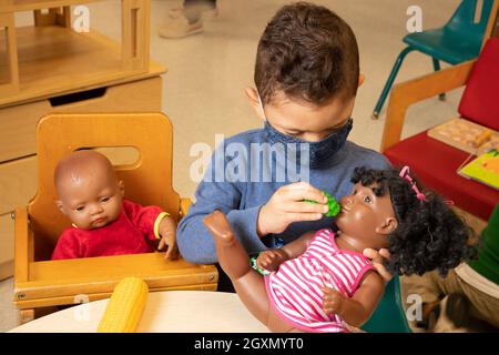 Istruzione Preschool 3-4 anni fingono di giocare nella zona della famiglia ragazzo che allatta bambola con cibo da gioco, indossare maschera viso a causa di Covid-19 Foto Stock