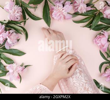 Due mani di una ragazza giovane con la pelle liscia e un mazzo di peonie rosa su uno sfondo di pesche, vista dall'alto, il concetto di moda per mano di cura della pelle, ant Foto Stock