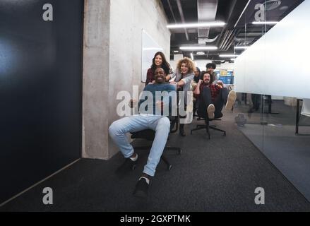 Multiethnics startup business team di sviluppatori di software per divertirsi durante la corsa su sedie da ufficio,entusiasti diversi dipendenti ridere godendo di un divertente Foto Stock