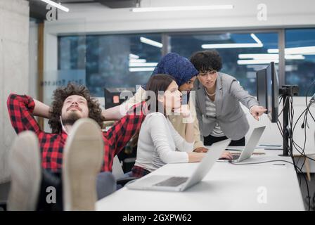 I giovani lo sviluppatore di software in appoggio con le gambe sulla scrivania mentre il suo multiethnics team aziendale la scrittura di codice di programmazione in background all'avvio di office Foto Stock