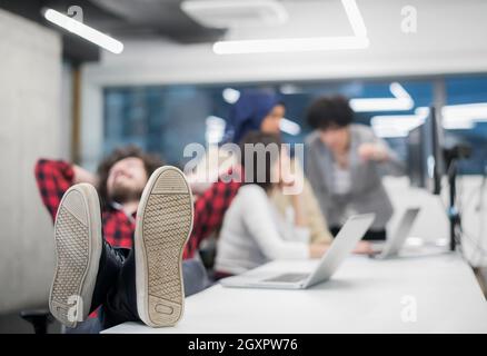 I giovani lo sviluppatore di software in appoggio con le gambe sulla scrivania mentre il suo multiethnics team aziendale la scrittura di codice di programmazione in background all'avvio di office Foto Stock