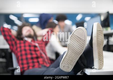 I giovani lo sviluppatore di software in appoggio con le gambe sulla scrivania mentre il suo multiethnics team aziendale la scrittura di codice di programmazione in background all'avvio di office Foto Stock