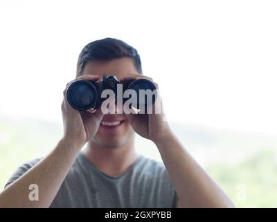 Giovane uomo che sta guardando attraverso una finestra di vetro con un binocolo come si guarda qualcosa di distanza Foto Stock