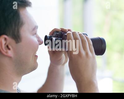 Giovane uomo che sta guardando attraverso una finestra di vetro con un binocolo come si guarda qualcosa di distanza Foto Stock