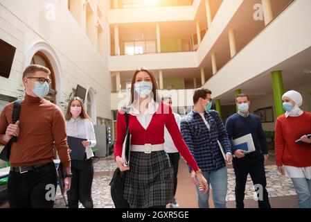 gruppo di studenti multietnici che camminano sul coridore universitario indossando maschera protettiva per il viso in un nuovo normale stile di vita pandemico del coronavirus nell'istruzione Foto Stock