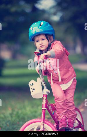 Carino sorridente bambina con la bicicletta e casco su strada nel parco Foto Stock