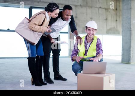 Ingegner architetto utilizzando computer portatile mentre mostra house design piani per una giovane coppia multietnica al sito in costruzione Foto Stock