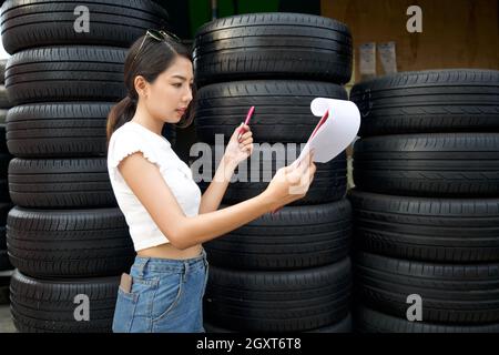 Il giovane proprietario asiatico del negozio di ricambi auto controlla il numero di pneumatici e gli ordini in arrivo prima di iniziare il lavoro al mattino. Diversi pneumatici auto sono Foto Stock
