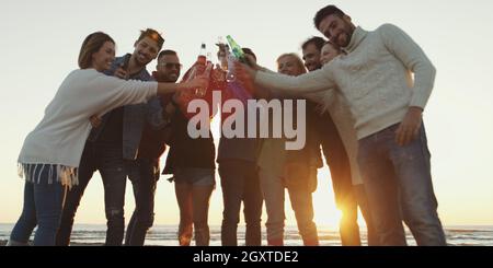 Giovani amici fare un brindisi con la birra e divertirsi intorno al falò presso il beach party sulla giornata autunnale Foto Stock