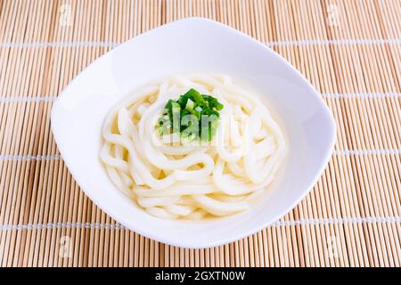 Tagliatelle di udon giapponesi preparate di fresco in ciotola bianca con cipolline guarnite su stuoia di bambù. Foto Stock