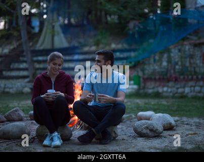 Coppia giovane seduto intorno al fuoco la sera tenendo un tazze di tè o caffè stessi di riscaldamento Foto Stock