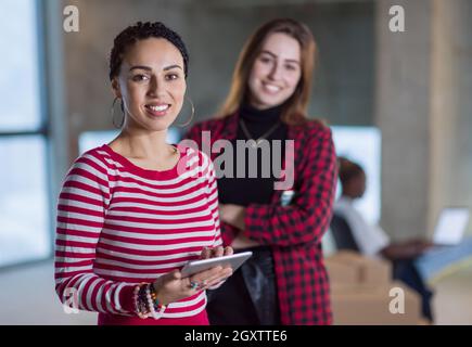 Due giovani donne d'affari allegre e casual con un collega afro-americano alle loro spalle che utilizzano un tablet sul cantiere durante il trasferimento in nuovi Foto Stock