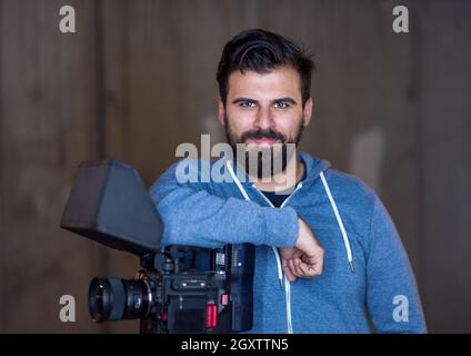 ritratto di un giovane videografo che si appoggia sulla fotocamera durante una pausa dalle riprese di fronte al muro di cemento Foto Stock