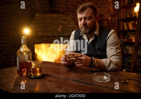 Uomo con ghigliottina taglia un sigaro, tavolo di legno su sfondo. Cultura del fumo di tabacco. Svaghi fumatore maschile con bicchiere di alcol Foto Stock