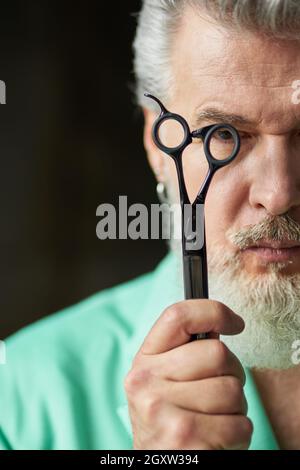 Ritratto corto di uomo maturo dai capelli grigi brutali con barba che guarda la fotocamera, tenendo forbici affilate vicino agli occhi, posando al buio Foto Stock