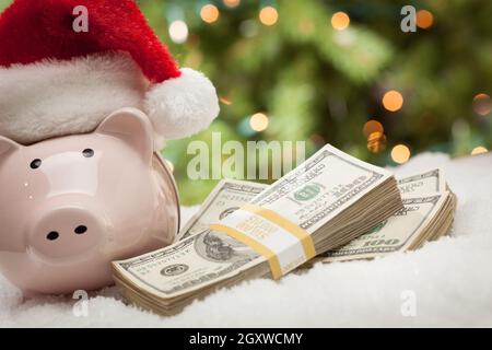 Salvadanaio indossando Santa Hat vicino a pile di centinaia di fatture del dollaro sui fiocchi di neve. Foto Stock