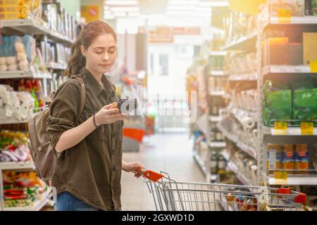 Una giovane bella donna rotola un carrello di alimentari e legge le informazioni sul prodotto selezionato. Concetto di consumo consapevole. Luce. Primo piano. Foto Stock