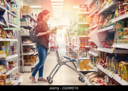 Una giovane donna bella rotola un carrello di alimentari e sceglie i prodotti nel supermercato. Luce. Foto Stock