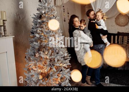 Famiglia gioiosa con i bambini in piedi vicino all'albero di Natale, festeggia le vacanze invernali insieme a casa. Bei genitori con figlio adorabile e. Foto Stock