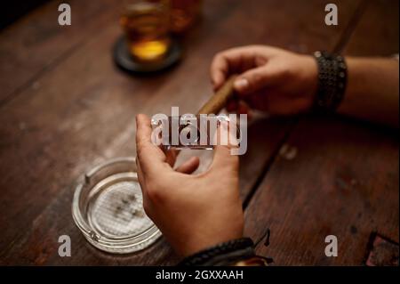 Uomo con ghigliottina taglia un sigaro, tavolo di legno su sfondo. Cultura del fumo di tabacco, sapore specifico. Svaghi fumatore maschile con bicchiere di alcol Foto Stock