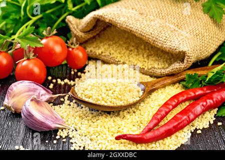 Couscous crudo in un cucchiaio e un sacco di burlap, pomodori, peperoni caldi, erbe e aglio su sfondo di legno Foto Stock