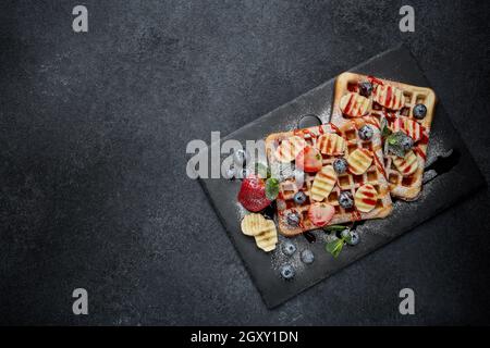 Deliziosi waffle con frutti di bosco, menta, zucchero in polvere e salsa dolce su tavola di ardesia su sfondo grigio. Vista dall'alto con spazio di copia. Pasto dolce. Foto Stock