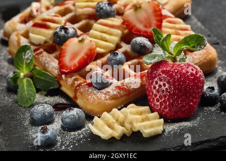 Deliziosi waffle con frutti di bosco, menta, zucchero in polvere e salsa dolce su tavola di ardesia su sfondo grigio. Pasto dolce. Dessert. Serve cibo Foto Stock