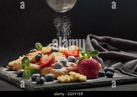 Deliziosi waffle con frutti di bosco, menta, zucchero a velo, salsa dolce su tavola di ardesia su sfondo scuro. Dessert. Serve cibo. Processo di sprink Foto Stock