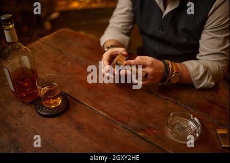 Uomo con ghigliottina taglia un sigaro, tavolo di legno su sfondo. Cultura del fumo di tabacco. Svaghi fumatore maschile con bicchiere di alcol Foto Stock