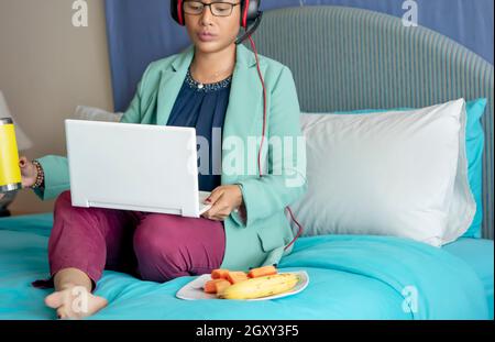 Donna d'affari o insegnante che indossa cuffie rosse sedute a letto e cibo in mano. Videoconferenza chiamata sul computer portatile parlare con zoom webcam t Foto Stock